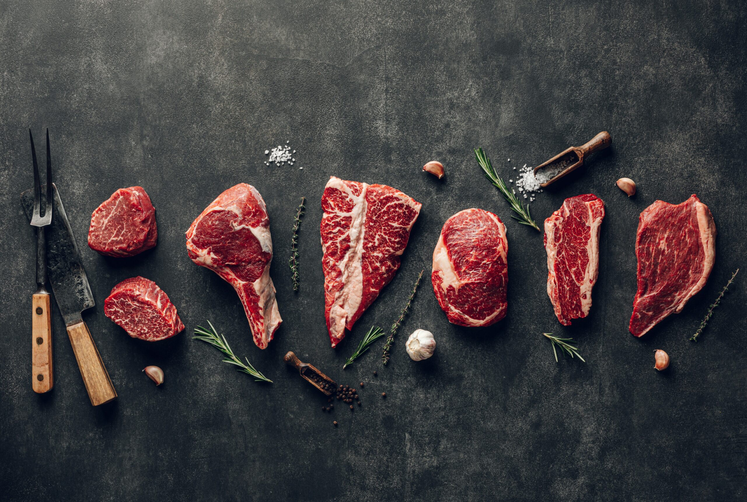 Steaks spread out across a grey background with herbs and utensils.